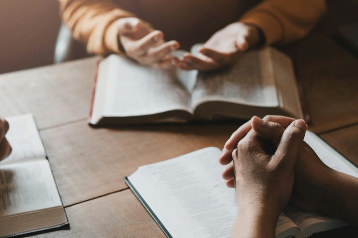 A group of Christians sit together and pray around a wooden table with blurred open Bible pages